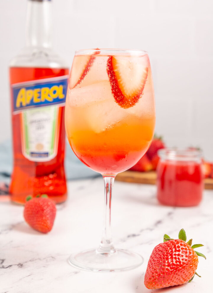 A strawberry aperol spritz in a wine glass next to a strawberry and bottle of Aperol.